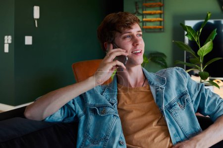 Téléchargez les photos : Young caucasian man wearing denim jacket smiling while using phone on sofa in living room at home - en image libre de droit