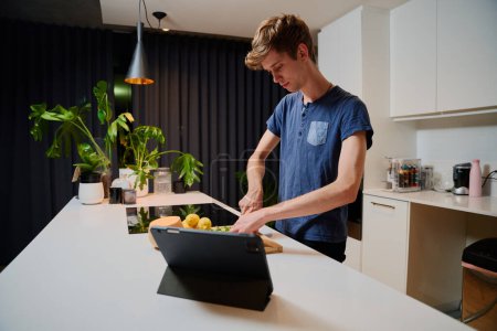 Téléchargez les photos : Jeune homme caucasien en vêtements décontractés préparer le dîner à côté de la tablette numérique dans la cuisine à la maison - en image libre de droit