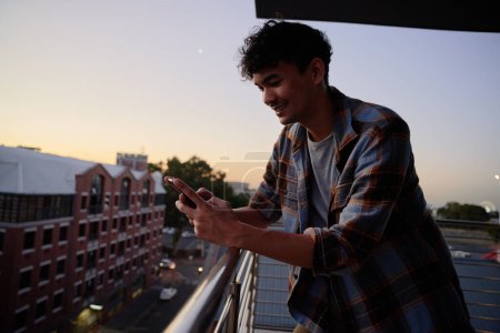Téléchargez les photos : Jeune homme multiracial en chemise à carreaux utilisant le téléphone sur le balcon de l'appartement pendant le coucher du soleil - en image libre de droit