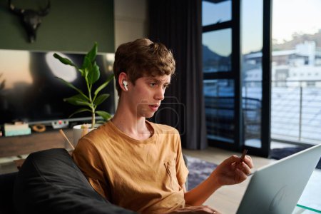 Téléchargez les photos : Jeune homme caucasien portant t-shirt tenant carte de crédit tout en faisant des achats en ligne sur ordinateur portable à la maison - en image libre de droit