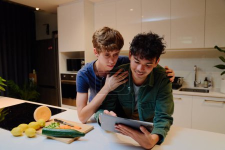 Photo for Young gay couple in casual clothing smiling while using digital tablet next to food in kitchen at home - Royalty Free Image