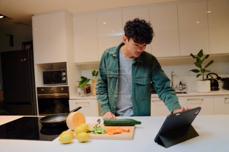 Téléchargez les photos : Jeune homme multiracial en chemise à manches longues utilisant une tablette numérique à côté de la nourriture dans la cuisine à la maison - en image libre de droit