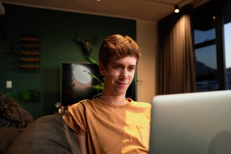 Téléchargez les photos : Jeune homme caucasien portant des vêtements décontractés souriant tout en utilisant un ordinateur portable sur le canapé dans le salon à la maison - en image libre de droit