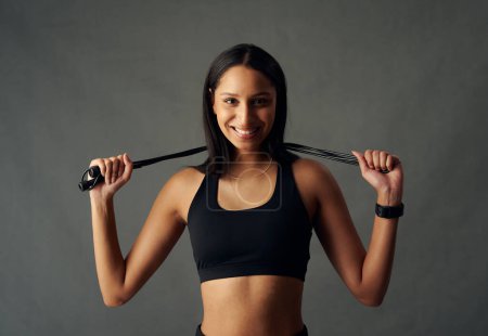 Photo for Portrait of happy young biracial woman wearing sports bra holding jump rope behind head in studio - Royalty Free Image