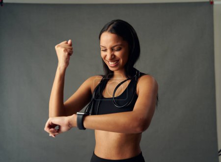 Foto de Happy young biracial woman wearing sports bra smiling while checking the time on fitness tracker in studio - Imagen libre de derechos