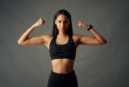 Foto de Mujer biracial joven con sujetador deportivo y rastreador de fitness flexionando los músculos del bíceps en el estudio - Imagen libre de derechos