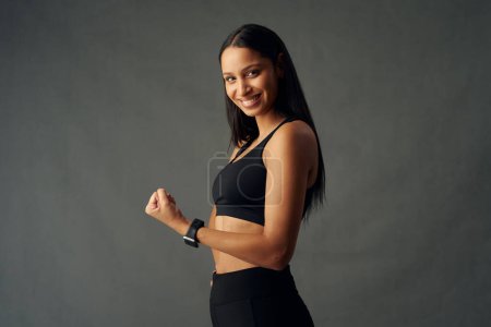 Téléchargez les photos : Young biracial woman wearing sports bra and fitness tracker smiling while flexing bicep muscles in studio - en image libre de droit