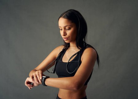 Téléchargez les photos : Focalisé jeune femme biraciale portant un soutien-gorge de sport regardant vers le bas à fitness tracker en studio - en image libre de droit