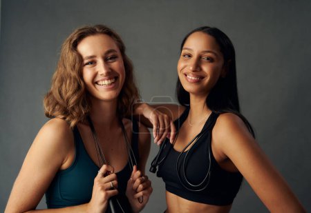 Foto de Young woman wearing sports bra holding jump rope while looking at camera and smiling with friend - Imagen libre de derechos