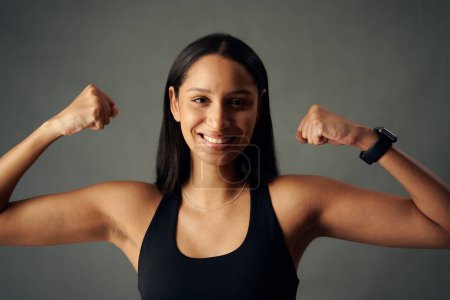 Photo for Portrait of happy young biracial woman wearing sports bra and fitness tracker flexing bicep muscles in studio - Royalty Free Image