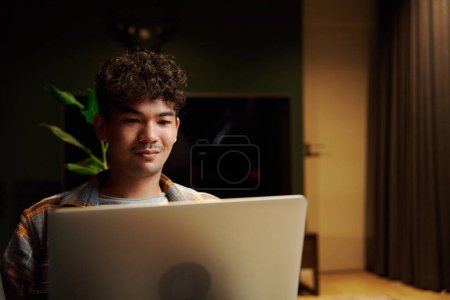 Téléchargez les photos : Young multiracial man wearing checkered shirt looking away while using laptop on sofa in house at night - en image libre de droit