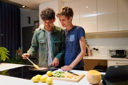 Photo for Young gay couple in casual clothing smiling while cooking dinner by digital tablet in kitchen at home - Royalty Free Image