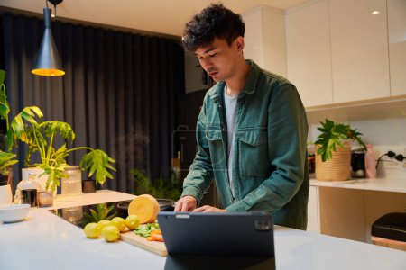 Photo for Young multiracial man wearing long sleeved shirt preparing dinner by digital tablet in kitchen at home - Royalty Free Image