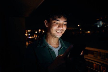 Photo for Young multiracial man wearing shirt smiling while using mobile phone on balcony of apartment at night - Royalty Free Image