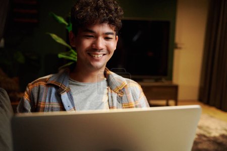 Téléchargez les photos : Heureux jeune homme multiracial portant chemise à carreaux souriant tout en utilisant un ordinateur portable sur le canapé à la maison - en image libre de droit