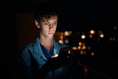 Téléchargez les photos : Focalisé jeune homme caucasien en vêtements décontractés textos sur téléphone portable à l'extérieur appartement la nuit - en image libre de droit