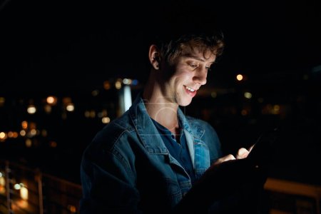 Téléchargez les photos : Jeune homme caucasien en vêtements décontractés souriant tout en tapant sur le téléphone mobile sur le balcon la nuit - en image libre de droit