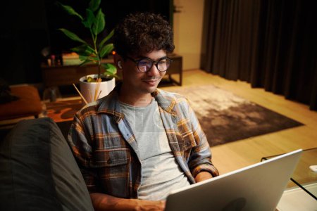 Foto de Young multiracial man wearing checkered shirt using laptop and wireless headphones on sofa at home - Imagen libre de derechos