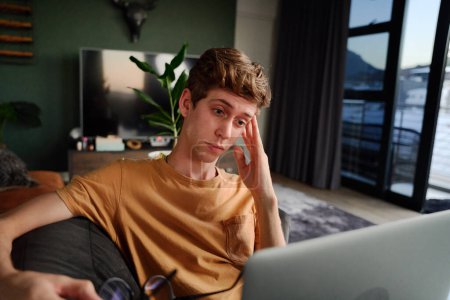 Foto de Exhausted young caucasian man holding eyeglasses next to laptop on sofa in living room at home - Imagen libre de derechos