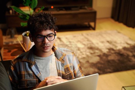 Foto de Focused young multiracial man wearing eyeglasses using laptop and wireless headphones on sofa at home - Imagen libre de derechos