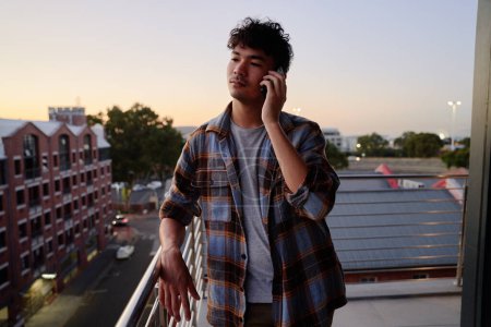 Téléchargez les photos : Jeune homme multiracial en chemise écoutant le téléphone mobile sur le balcon de l'appartement pendant le coucher du soleil - en image libre de droit