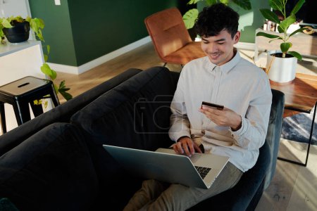 Téléchargez les photos : Jeune homme multiracial souriant tout en utilisant un ordinateur portable et carte de crédit sur le canapé dans le salon à la maison - en image libre de droit