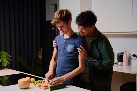 Foto de Joven pareja gay en ropa casual sonriendo y abrazando mientras prepara la cena en la cocina en casa - Imagen libre de derechos