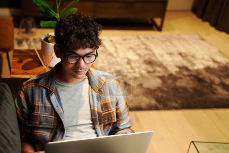 Téléchargez les photos : Focalisé jeune homme multiracial portant des lunettes et chemise à carreaux à l'aide d'un ordinateur portable sur le canapé à la maison - en image libre de droit