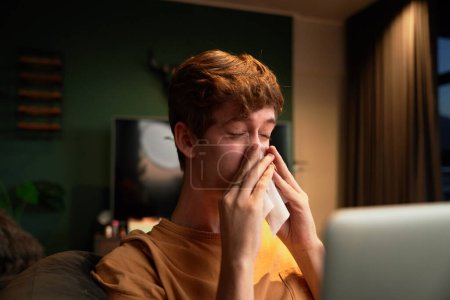 Téléchargez les photos : Malade jeune homme caucasien en vêtements décontractés se mouchant à côté de l'ordinateur portable sur le canapé dans le salon à la maison - en image libre de droit