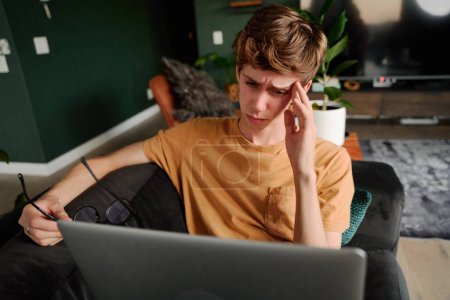 Téléchargez les photos : Frustré jeune homme caucasien en vêtements décontractés tenant des lunettes par ordinateur portable sur canapé dans le salon à la maison - en image libre de droit