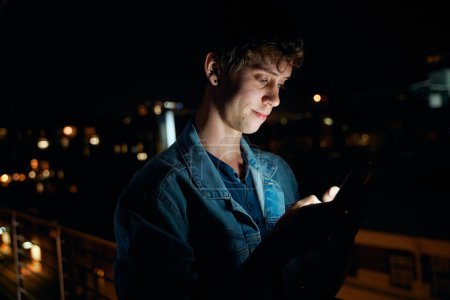 Téléchargez les photos : Jeune homme caucasien en vêtements décontractés textos sur téléphone portable à l'extérieur appartement la nuit - en image libre de droit