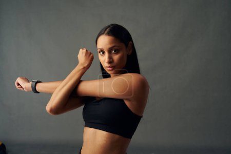 Photo for Young biracial woman wearing sports bra looking at camera while doing cross arm stretch in studio - Royalty Free Image