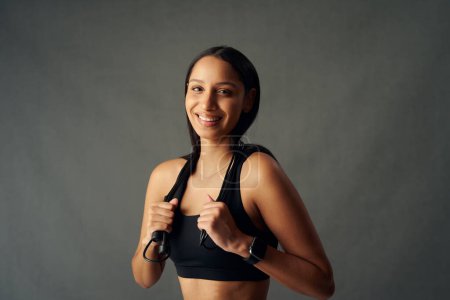 Foto de Portrait of happy young biracial woman wearing sports bra holding jump rope over shoulders in studio - Imagen libre de derechos