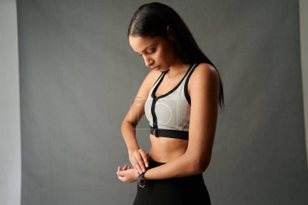 Foto de Mujer biracial joven en ropa deportiva ajustando el reloj de seguimiento de fitness frente al telón de fondo en el estudio - Imagen libre de derechos