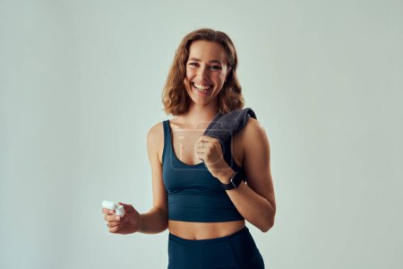 Foto de Feliz joven mujer caucásica usando ropa deportiva sosteniendo auriculares inalámbricos y toalla en el estudio - Imagen libre de derechos