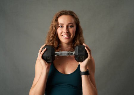 Photo for Portrait of young caucasian woman wearing sports bra smiling while holding dumbbell in studio - Royalty Free Image