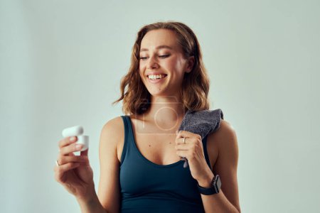 Foto de Mujer caucásica joven usando ropa deportiva sonriendo mientras sostiene auriculares inalámbricos y toalla en el estudio - Imagen libre de derechos