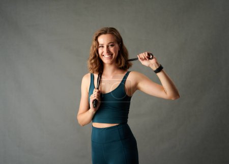 Téléchargez les photos : Trois quarts de longueur de jeune femme caucasienne en vêtements de sport souriant tout en tenant corde à sauter en studio - en image libre de droit