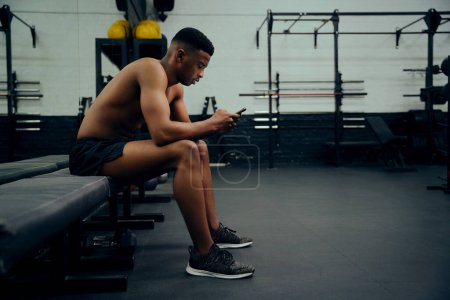 Téléchargez les photos : Jeune homme afro-américain regardant son téléphone pour répondre à un message texte. Mixte, entraîneur personnel masculin regardant son téléphone tout en s'asseyant dans la salle de gym. Photo de haute qualité - en image libre de droit