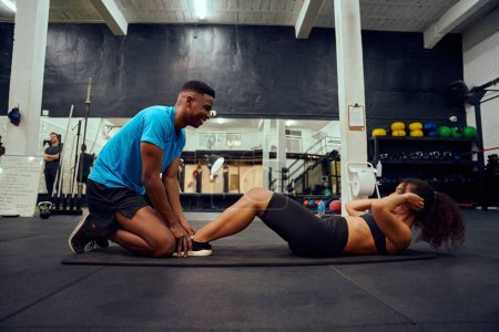 Foto de Multi-ethnic friends doing cross fit in the gym. African American male encouraging African American female during sit-ups. High quality photo - Imagen libre de derechos