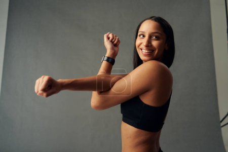 Téléchargez les photos : Young biracial woman wearing sports bra looking away and smiling while doing cross arm stretch in studio - en image libre de droit