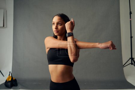 Foto de Focused young biracial woman wearing sports clothing and fitness tracker doing cross arm stretch in studio - Imagen libre de derechos