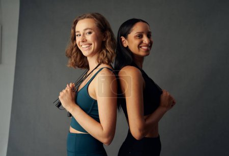 Foto de Mujeres jóvenes que usan ropa deportiva de espalda a espalda mientras sonríen y sostienen la cuerda de salto - Imagen libre de derechos
