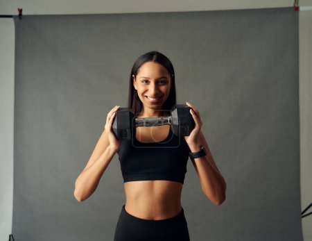 Photo for Happy young biracial woman wearing sports bra smiling while holding dumbbell in studio - Royalty Free Image