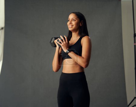 Téléchargez les photos : Happy young biracial woman wearing sports bra and leggings looking away while holding weights in studio - en image libre de droit