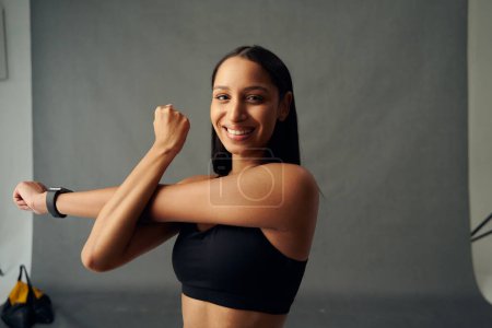 Téléchargez les photos : Jeune femme biraciale en vêtements de sport souriant tout en faisant l'étirement du bras croisé et en regardant la caméra en studio - en image libre de droit