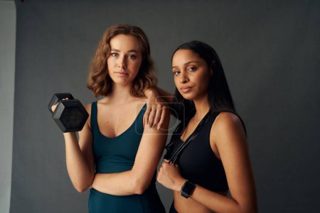 Photo for Young woman with friend wearing sports clothing looking at camera while holding dumbbell - Royalty Free Image