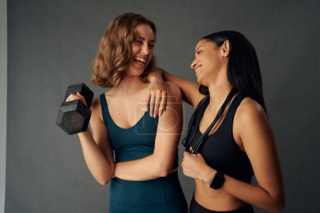 Téléchargez les photos : Jeunes femmes portant des vêtements de sport riant tandis que face à face tenant haltère - en image libre de droit
