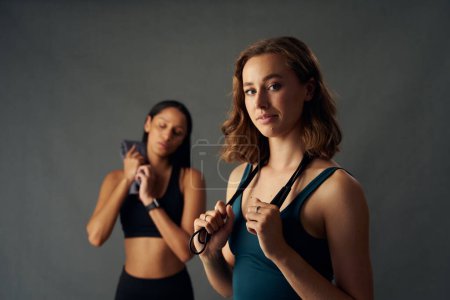 Foto de Mujeres jóvenes seguras que usan ropa deportiva sosteniendo la cuerda de salto sobre el hombro mientras miran a la cámara - Imagen libre de derechos