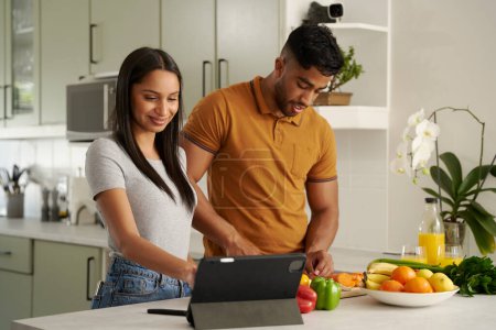 Téléchargez les photos : Jeune couple biracial portant des vêtements décontractés préparant la nourriture à côté de la tablette numérique dans la cuisine - en image libre de droit